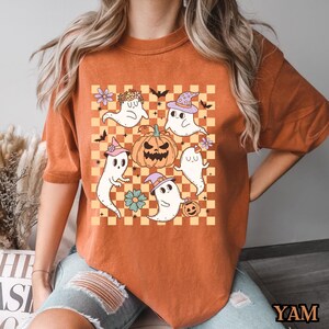 May include: Orange t-shirt with a checkered pattern featuring a group of cute ghosts and a jack-o-lantern. The ghosts are wearing hats and holding pumpkins.