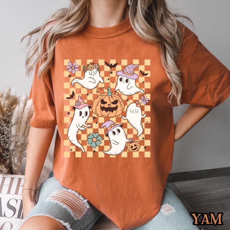 May include: Orange t-shirt with a checkered pattern featuring a group of cute ghosts and a jack-o-lantern. The ghosts are wearing hats and holding pumpkins.