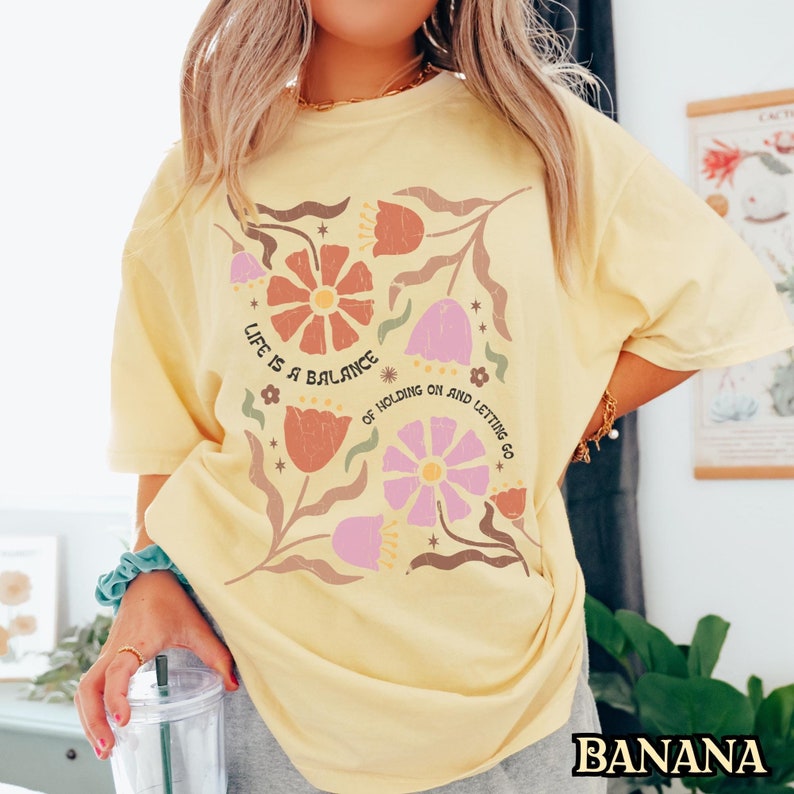 Pu&ograve; includere: Una maglietta gialla con un disegno floreale e il testo "Life is a balance of holding on and letting go".