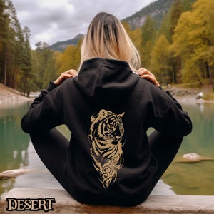 May include: Black hoodie with a gold tiger graphic on the back. The tiger is facing to the right and has a detailed design. The hoodie is worn by a person sitting on a rock by a lake.