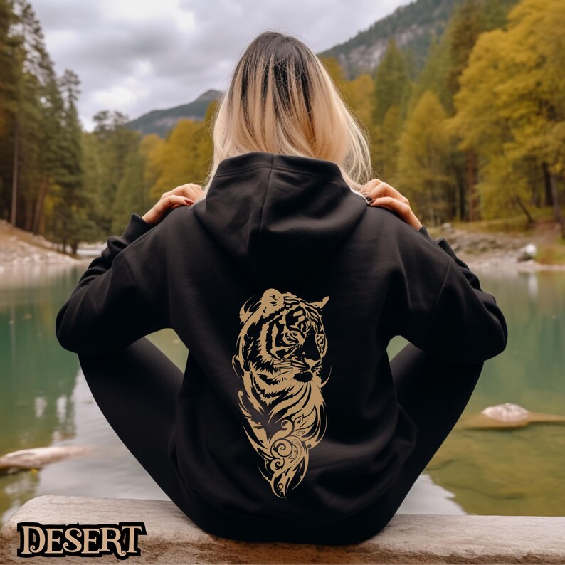 May include: Black hoodie with a gold tiger graphic on the back. The tiger is facing to the right and has a detailed design. The hoodie is worn by a person sitting on a rock by a lake.