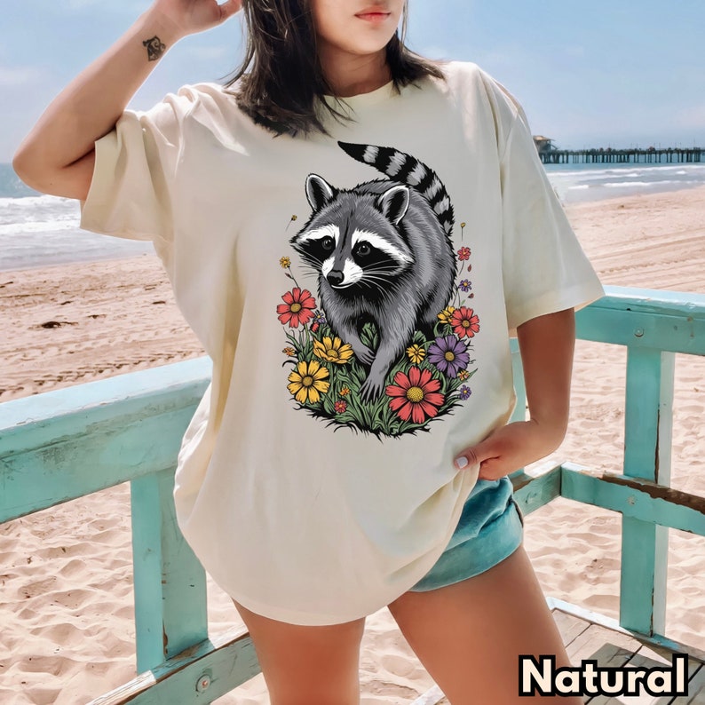 May include: A cream colored t-shirt with a graphic of a raccoon surrounded by colorful flowers. The text "Natural" is visible at the bottom of the image.