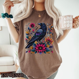 May include: A brown t-shirt with a black raven and colorful floral design. The raven is perched on a branch with flowers in shades of pink, purple, yellow, and green.