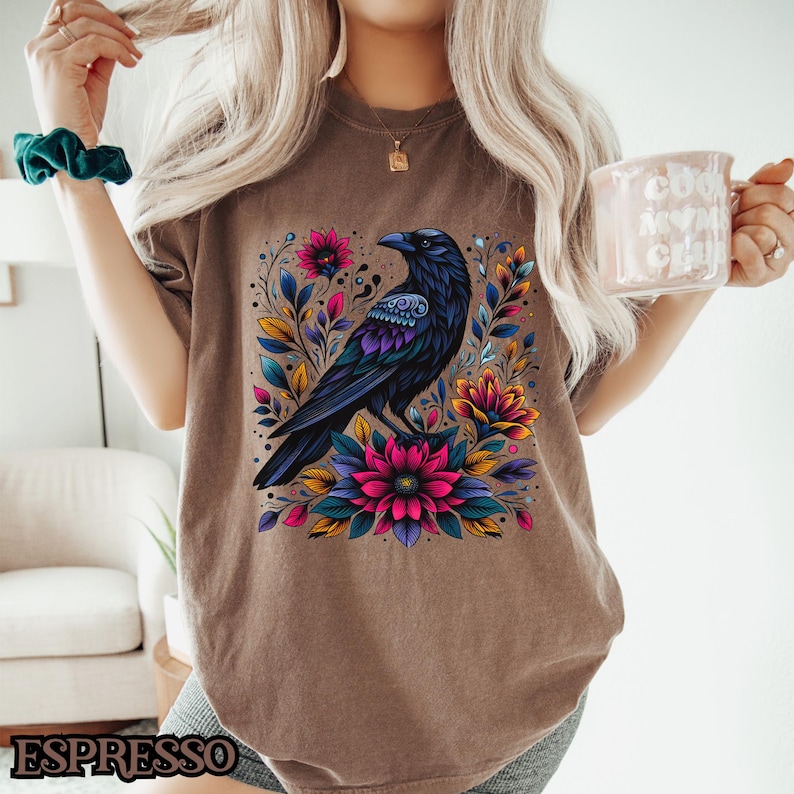 May include: A brown t-shirt with a black raven and colorful floral design. The raven is perched on a branch with flowers in shades of pink, purple, yellow, and green.