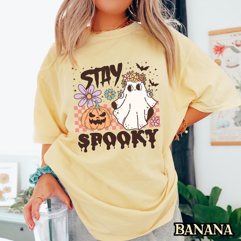 K&ouml;nnte beinhalten: Ein gelbes T-Shirt mit einem grafischen Aufdruck eines Geistes, der eine Blumenkrone tr&auml;gt, einen K&uuml;rbis und den Text "Stay Spooky".