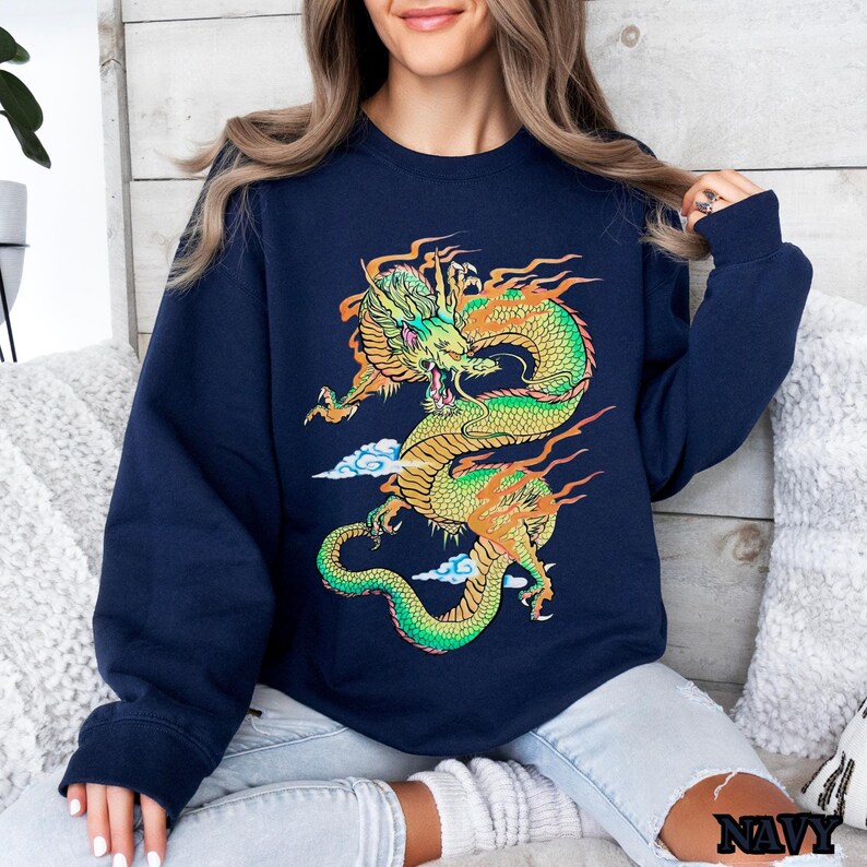 K&ouml;nnte beinhalten: Ein marineblauer Pullover mit einem bunten Drachen-Motiv. Der Drache ist gr&uuml;n, gelb und orange und hat ein Wolkenmuster dahinter.