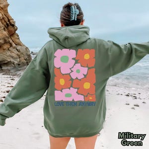 May include: A military green hooded sweatshirt featuring a large graphic on the back with stylized flowers in shades of pink, orange, and yellow. The text "LOVE THEM ANYWAY" is printed in blue letters beneath the floral design on the back.
