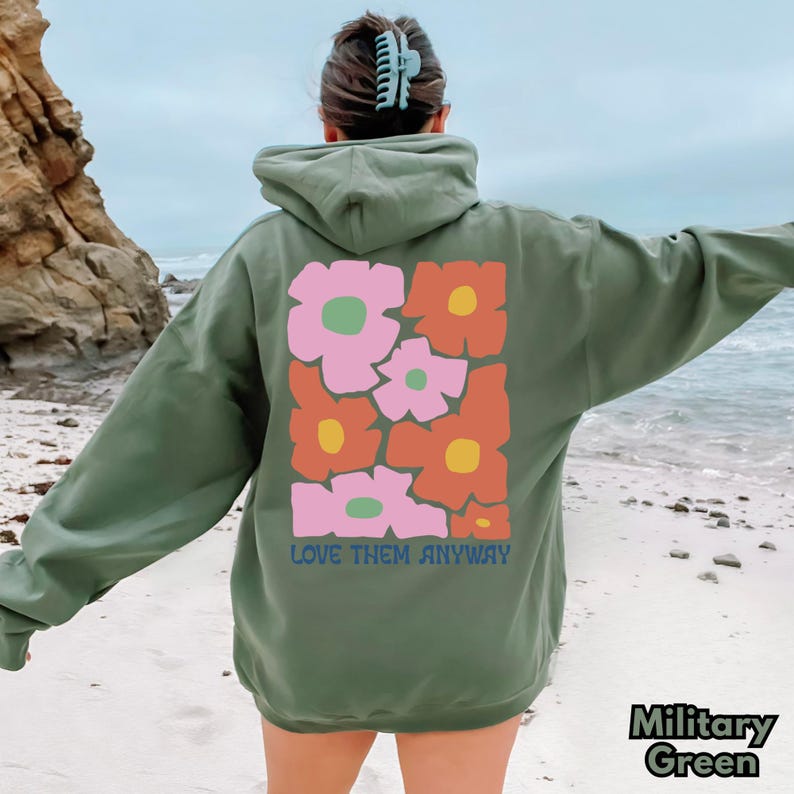 May include: A military green hooded sweatshirt featuring a large graphic on the back with stylized flowers in shades of pink, orange, and yellow. The text "LOVE THEM ANYWAY" is printed in blue letters beneath the floral design on the back.