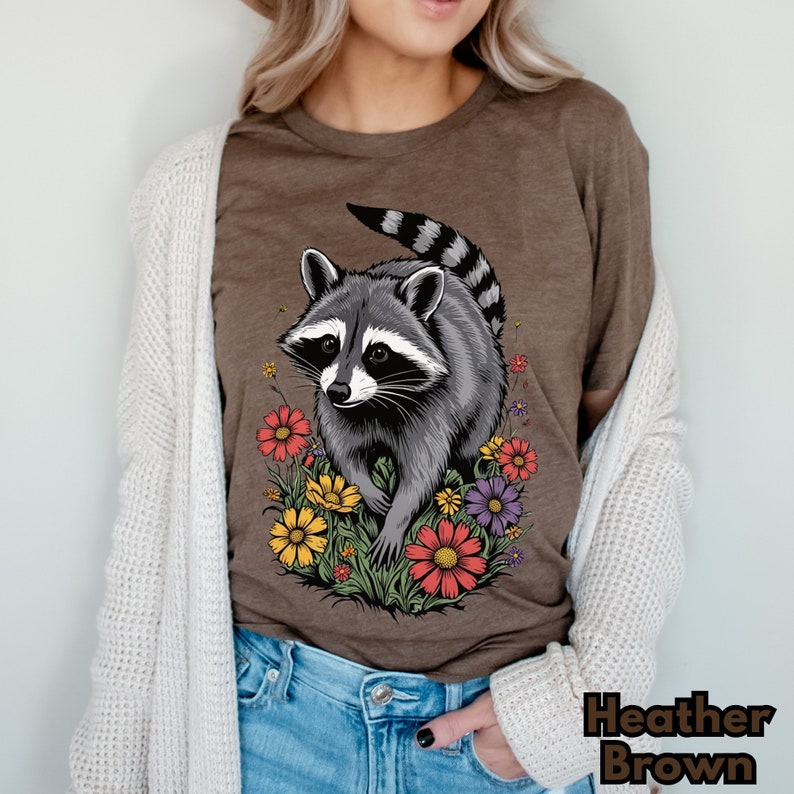 May include: A brown t-shirt with a graphic of a raccoon walking through a field of colorful flowers. The raccoon is gray and black with a white mask. The flowers are red, yellow, purple, and orange.