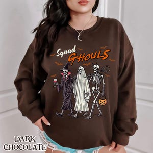 Pu&ograve; includere: Una felpa marrone con una stampa grafica di tre scheletri vestiti da strega, fantasma e scheletro. Il testo "Squad Ghouls" &egrave; stampato sopra gli scheletri.