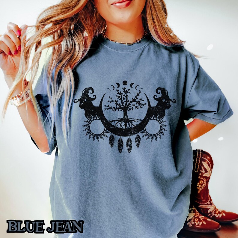 May include: A blue t-shirt with a black graphic of a tree with roots, a crescent moon, and two rams' heads. The graphic is surrounded by a circle of small moons.