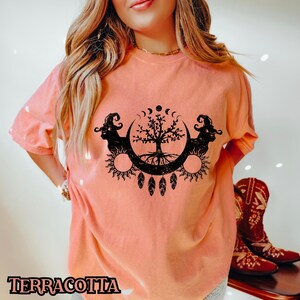 May include: A terracotta colored t-shirt with a black graphic of a tree with roots, surrounded by a crescent moon and sun. The tree is flanked by two black figures with horns and a witch's hat.