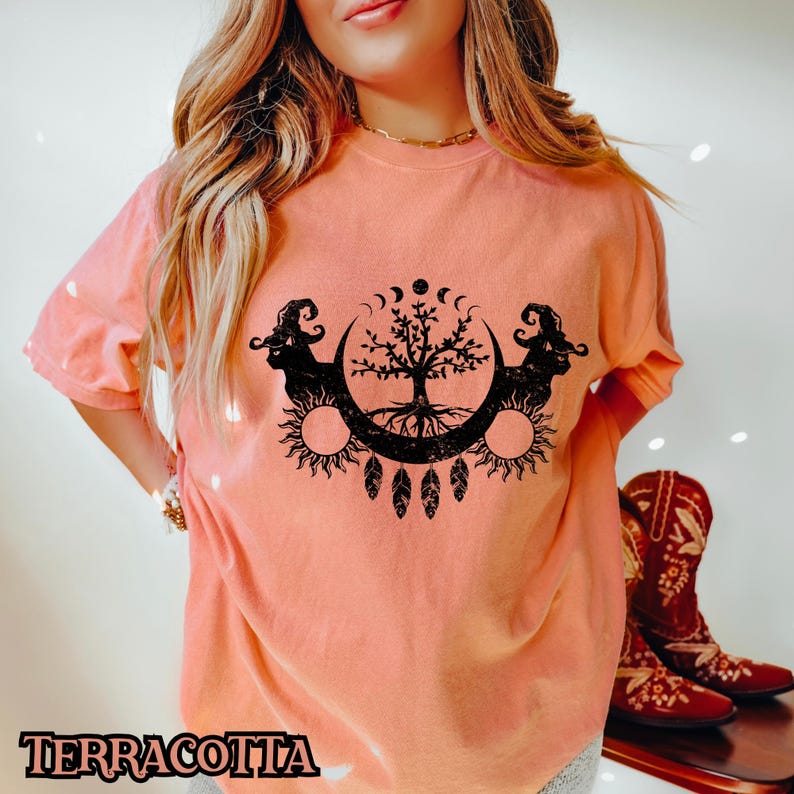 May include: A terracotta colored t-shirt with a black graphic of a tree with roots, surrounded by a crescent moon and sun. The tree is flanked by two black figures with horns and a witch's hat.