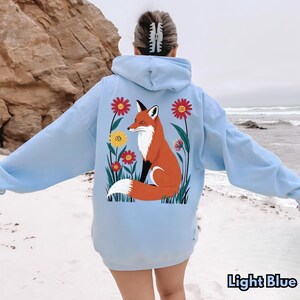 May include: Light blue hoodie with a fox illustration. The back of the hoodie features a vibrant orange fox with a white chest and tail, surrounded by red and yellow flowers and green foliage. The text "Light Blue" is at the bottom.