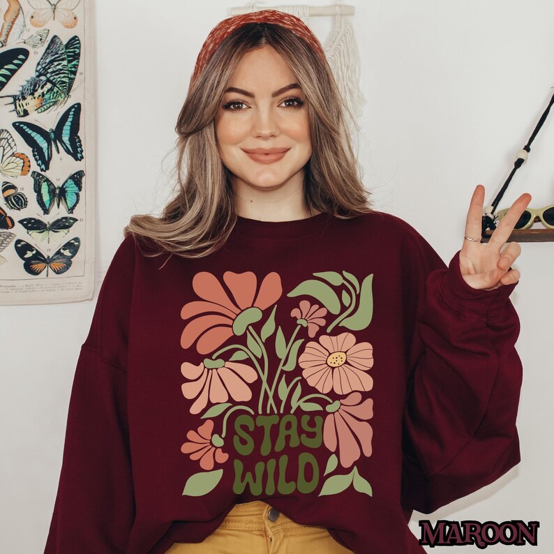 May include: A maroon sweatshirt featuring a floral design with the text "STAY WILD". The flowers are in shades of pink and peach with green leaves. The word "MAROON" is printed in the bottom right corner.