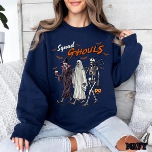 Pu&ograve; includere: Una felpa blu navy con una grafica di tre scheletri vestiti per Halloween. Il testo "Squad Ghouls" &egrave; stampato sulla felpa.