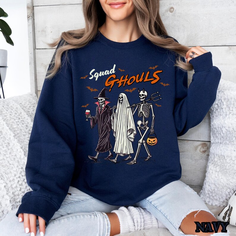 Pu&ograve; includere: Una felpa blu navy con una grafica di tre scheletri vestiti per Halloween. Il testo "Squad Ghouls" &egrave; stampato sulla felpa.