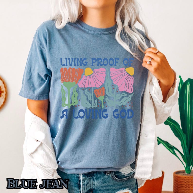 May include: A blue jean t-shirt with a graphic of pink, red, and yellow flowers and the text "Living Proof of a Loving God".