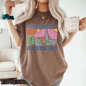 May include: A brown t-shirt with a graphic of colorful flowers and the text "Living Proof of a Loving God".