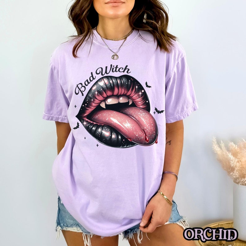 以下が含まれることがあります： ラベンダー色のTシャツに、黒とピンクのグリッターでできた唇のグラフィックがプリントされています。舌が突き出ていて、牙があります。唇の上に「Bad Witch」という文字があります。