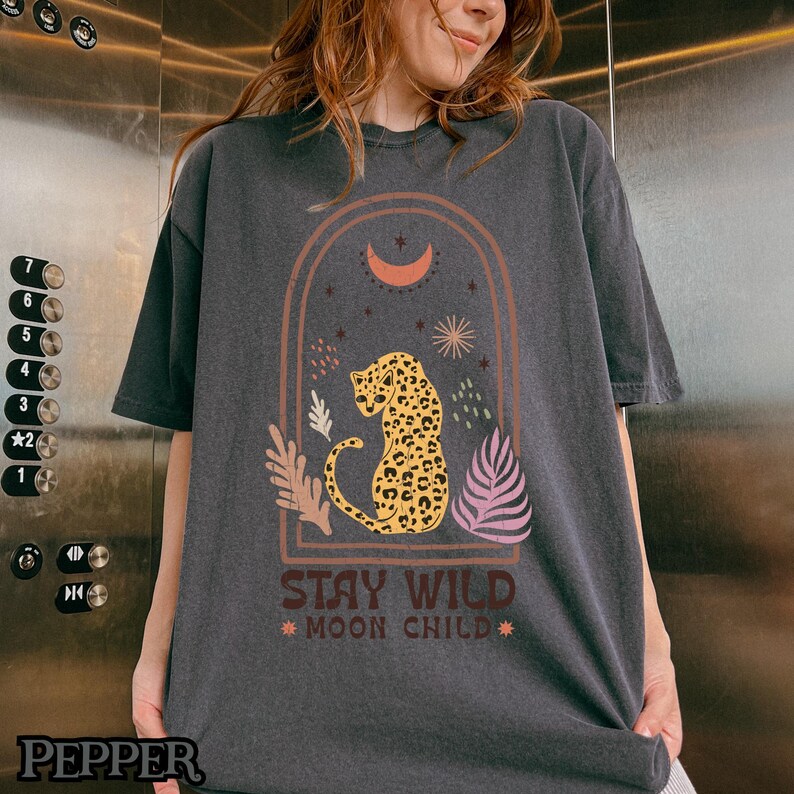 May include: A charcoal gray short-sleeve t-shirt featuring a graphic print of a leopard inside an arched frame. The design includes a crescent moon, stars, and botanical elements with the text STAY WILD MOON CHILD printed below the leopard.