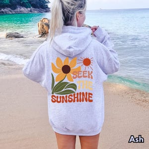 K&ouml;nnte beinhalten: Ein grauer Hoodie mit einer Sonnenblume und dem Text "Seek the Sunshine" auf der R&uuml;ckseite. Der Hoodie wird von einer Person getragen, die an einem Strand steht.