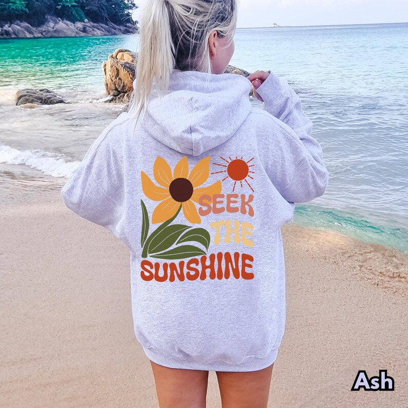 K&ouml;nnte beinhalten: Ein grauer Hoodie mit einer Sonnenblume und dem Text "Seek the Sunshine" auf der R&uuml;ckseite. Der Hoodie wird von einer Person getragen, die an einem Strand steht.