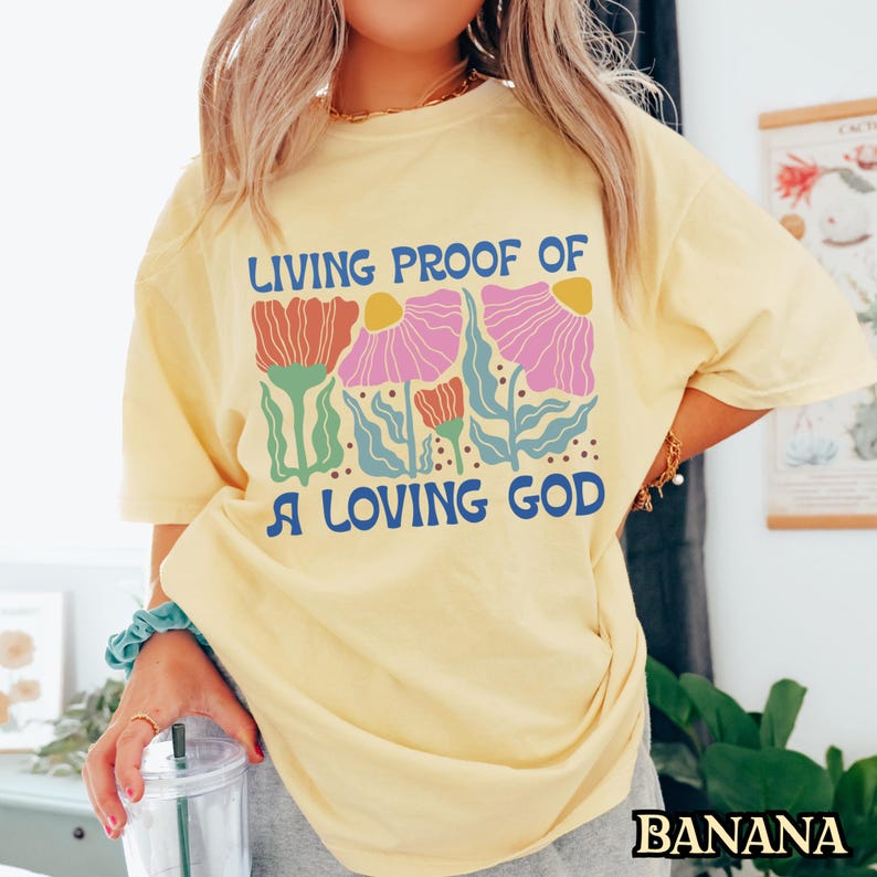 May include: A yellow t-shirt with a floral design and the text "Living Proof of a Loving God".