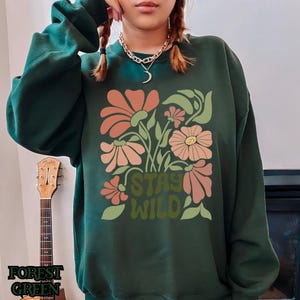 May include: Dark green sweatshirt featuring a floral design in peach and green hues. The words "STAY WILD" are printed in green. A guitar is visible in the background.