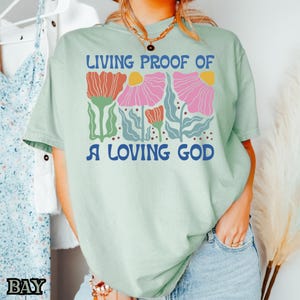 May include: A light green t-shirt with a floral design and the text "Living Proof of a Loving God".