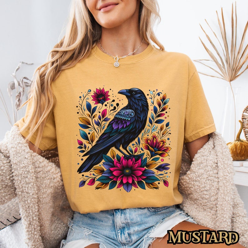 May include: A mustard yellow t-shirt with a black crow surrounded by colorful flowers. The crow has blue and purple accents on its wings and body.