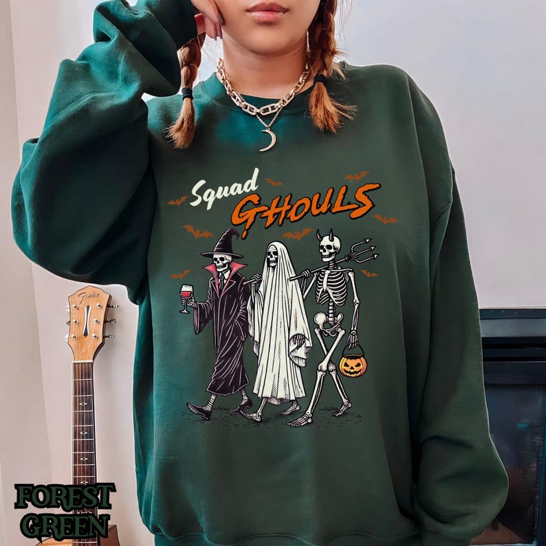 Pu&ograve; includere: Una felpa verde scuro con una stampa grafica di tre scheletri vestiti per Halloween. Il testo "Squad Ghouls" &egrave; stampato sopra gli scheletri.