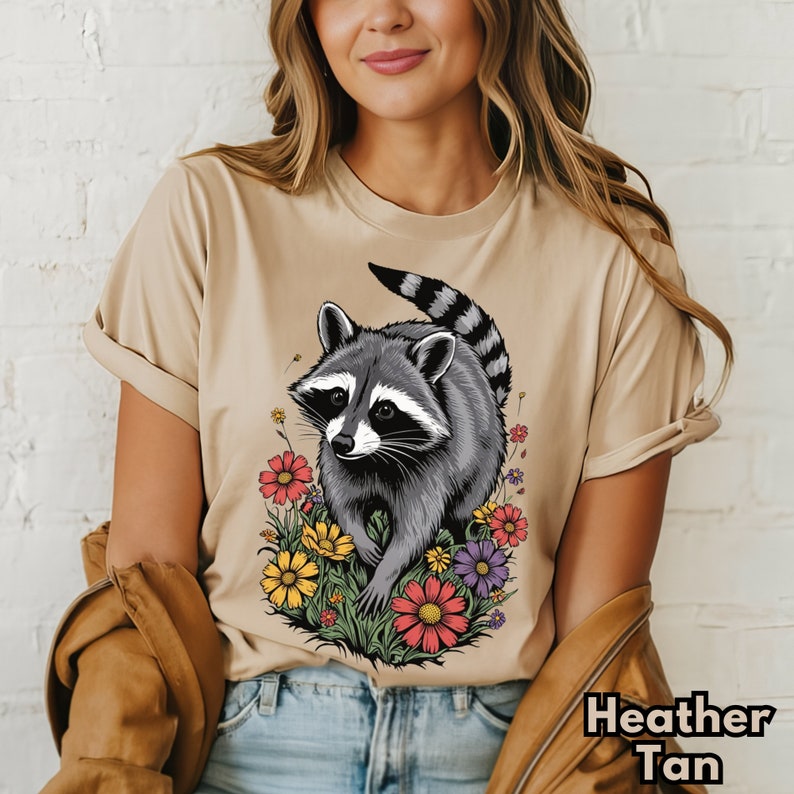 May include: A tan t-shirt with a graphic of a raccoon surrounded by colorful flowers. The text "Heather Tan" is visible in the bottom right corner of the image.