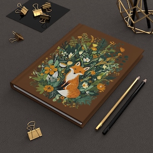 Può includere: Un diario rilegato in pelle marrone con una volpe circondata da fiori e vegetazione. La volpe è arancione e bianca con una coda folta. I fiori sono gialli, bianchi e rosa.