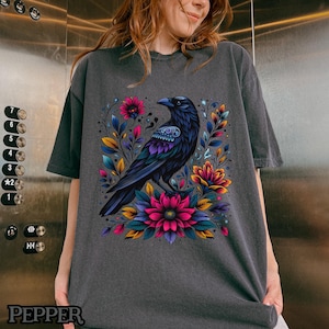 May include: A dark gray t-shirt with a black raven and colorful floral design. The raven is perched on a branch with flowers in shades of pink, purple, and yellow.