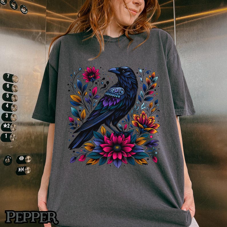 May include: A dark gray t-shirt with a black raven and colorful floral design. The raven is perched on a branch with flowers in shades of pink, purple, and yellow.