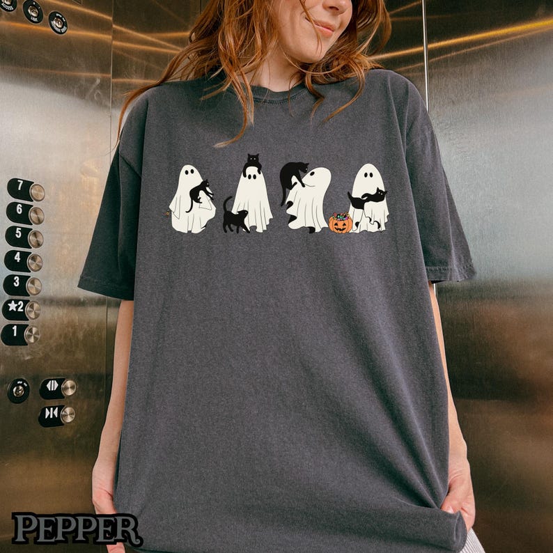 K&ouml;nnte beinhalten: Ein dunkelgraues T-Shirt mit einem Aufdruck von f&uuml;nf Geistern, die schwarze Katzen halten. Die Geister sind wei&szlig; und haben schwarze Augen. Die Katzen sind schwarz und haben wei&szlig;e Augen. Die Geister halten die Katzen in ihren Armen. Vor den Geistern befindet sich ein kleiner orangefarbener K&uuml;rbis.