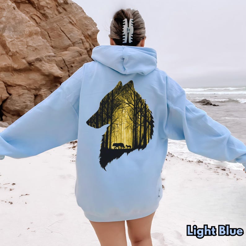 May include: A light blue hooded sweatshirt featuring a large graphic on the back. The graphic is shaped like a wolf head silhouette and contains a detailed illustration of a forest with tall trees and two wolves walking in the yellow light.