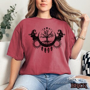 May include: A red t-shirt with a black graphic of a tree of life with crescent moons, suns, and feathers. The tree is surrounded by two rams' heads.