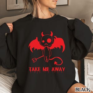 以下が含まれることがあります： 黒いスウェットシャツ。赤い悪魔のグラフィック、角、翼、三叉の槍が含まれています。「TAKE ME AWAY」の文字がグラフィックの下に赤で印刷されています。「BLACK」の文字が右下に印刷されています。