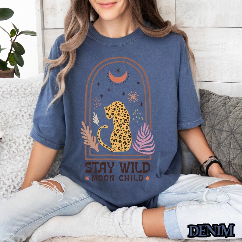 May include: A denim blue short-sleeve t-shirt featuring a graphic print of a leopard inside an arched frame with a crescent moon, stars, and foliage. The text "STAY WILD MOON CHILD" is printed in brown below the graphic. The shirt has a crew neckline.