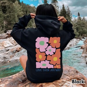 May include: A black hooded sweatshirt featuring a large rectangular graphic on the back. The design consists of stylized, blocky flowers in shades of pink, orange, and yellow with green centers. The text "LOVE THEM ANYWAY" is printed below.
