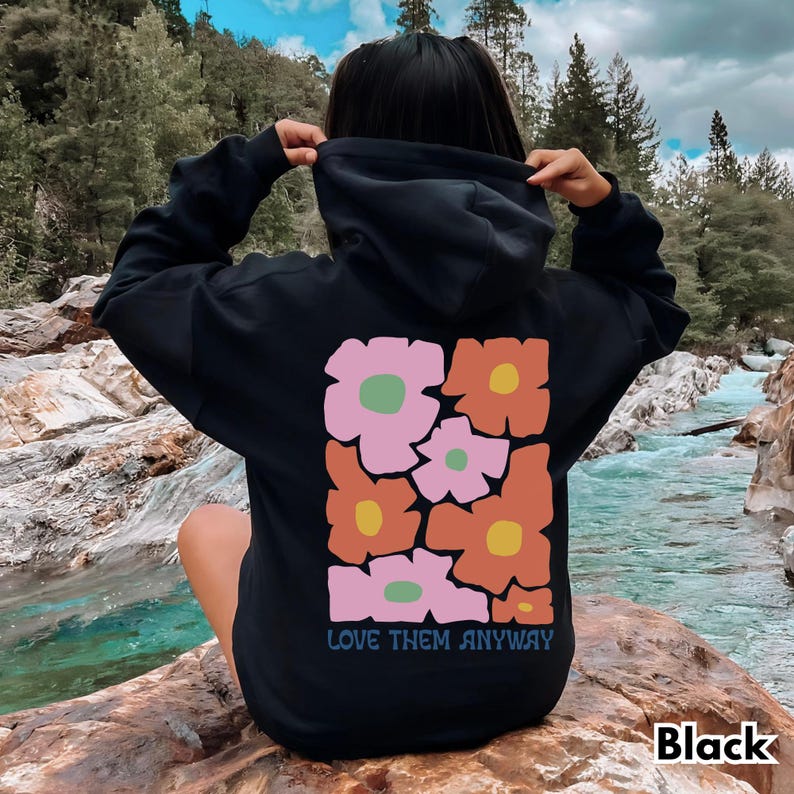 May include: A black hooded sweatshirt featuring a large rectangular graphic on the back. The design consists of stylized, blocky flowers in shades of pink, orange, and yellow with green centers. The text "LOVE THEM ANYWAY" is printed below.