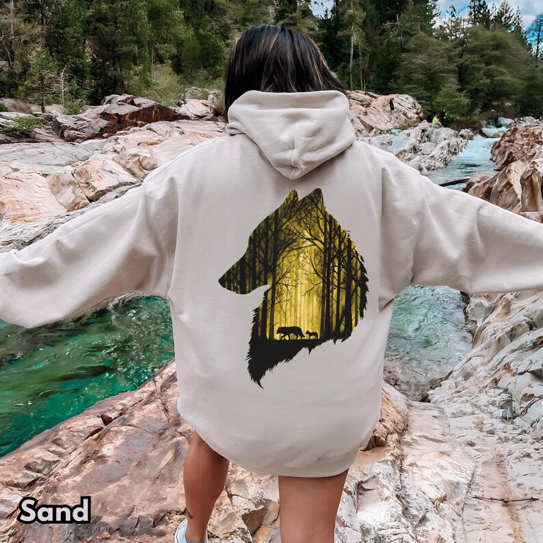 May include: A sand-colored hooded sweatshirt featuring a large graphic on the back. The graphic is a silhouette of a wolf head filled with a yellow-toned forest scene, including tall trees and two smaller wolf figures walking within the woods.