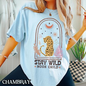 May include: A light blue t-shirt featuring a graphic of a leopard sitting inside an arched frame. The design includes a crescent moon, stars, and foliage elements. Below the graphic, the text reads STAY WILD MOON CHILD in dark brown letters.