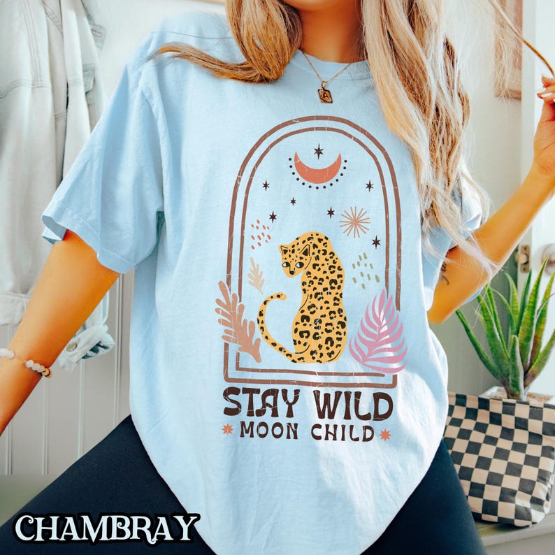 May include: A light blue t-shirt featuring a graphic of a leopard sitting inside an arched frame. The design includes a crescent moon, stars, and foliage elements. Below the graphic, the text reads STAY WILD MOON CHILD in dark brown letters.