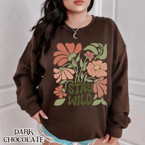 May include: Dark chocolate-coloured sweatshirt with a floral design. It features orange, peach, and yellow flowers, green leaves, and the words "STAY WILD" in green. The words "DARK CHOCOLATE" are at the bottom.