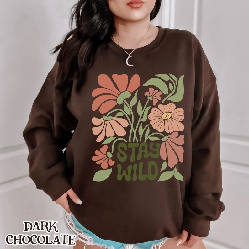 May include: Dark chocolate-coloured sweatshirt with a floral design. It features orange, peach, and yellow flowers, green leaves, and the words "STAY WILD" in green. The words "DARK CHOCOLATE" are at the bottom.