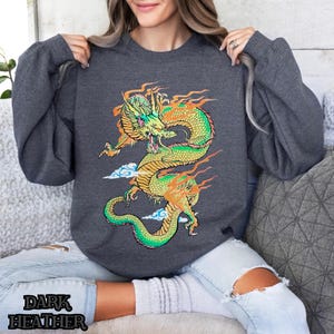K&ouml;nnte beinhalten: Ein dunkel-heathergrauer Sweatshirt mit einem grafischen Aufdruck eines gr&uuml;nen und gelben Drachen mit orangefarbenen Flammen. Der Drache ist von wei&szlig;en Wolken umgeben.