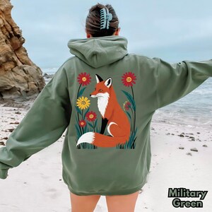 May include: Military green hoodie with a fox and floral graphic on the back. The fox is orange and white, surrounded by red and yellow flowers. The hoodie has a hood and long sleeves. The text "Military Green" is at the bottom right.